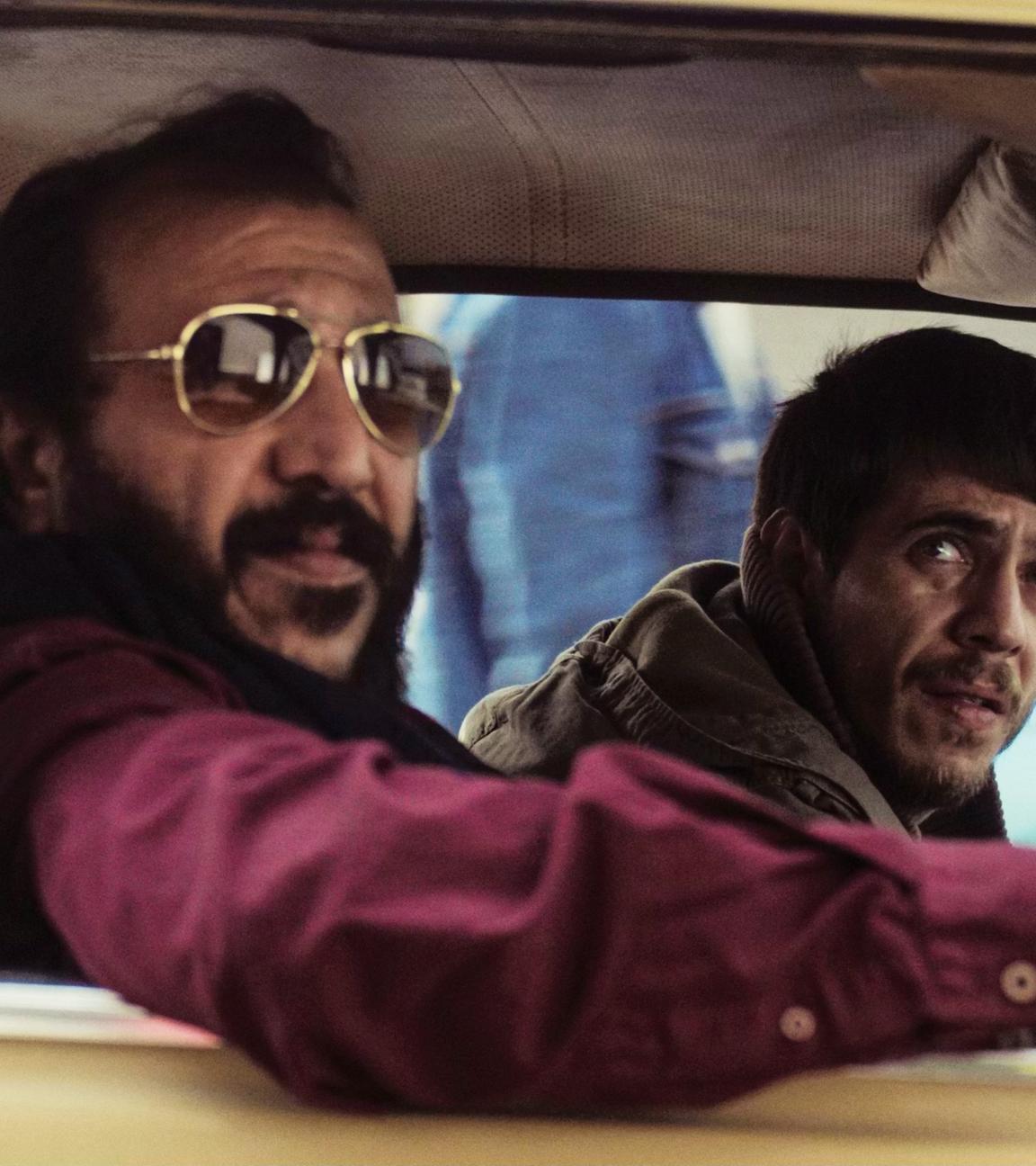 Zwei dunkelhaarige Männer, der eine mit Bart und Sonnenbrille, sitzen im Auto und schauen aus dem Fenster.