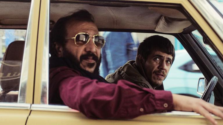 Zwei dunkelhaarige Männer, der eine mit Bart und Sonnenbrille, sitzen im Auto und schauen aus dem Fenster.
