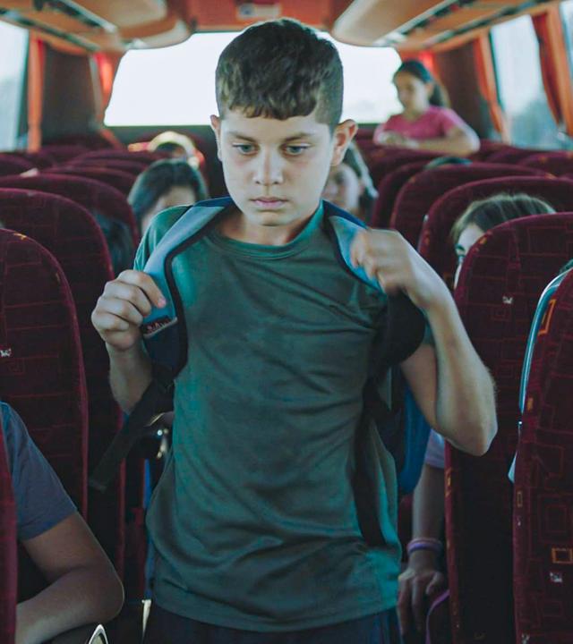 Ein junge mit Schulranzen auf dem Rücken geht durch den Gang zwischen den Sitzreihen in einem Bus.