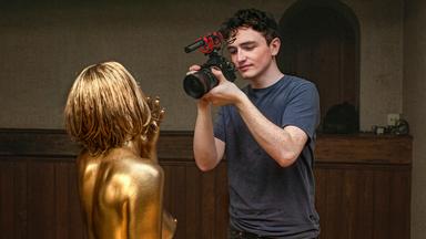 Rechts im Bild steht ein junger Mann, der eine Videokamera in der Hand hält und eine Frau filmt, deren Haare und Körper gold angemalt sind.