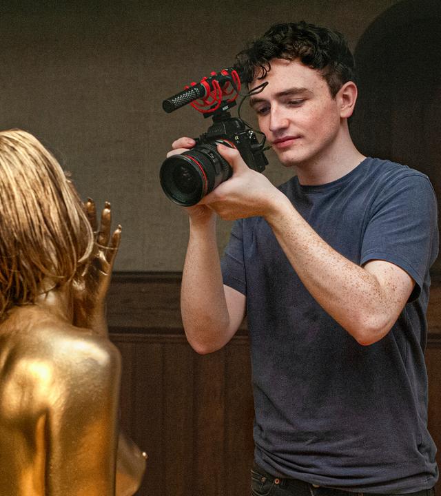 Rechts im Bild steht ein junger Mann, der eine Videokamera in der Hand hält und eine Frau filmt, deren Haare und Körper gold angemalt sind.