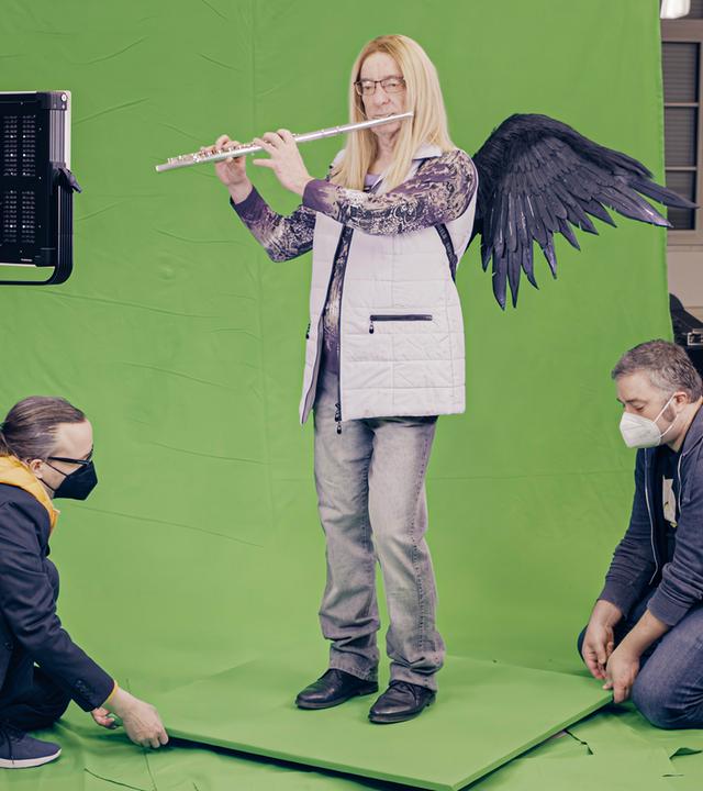 Eine Flötistin Mit umgebundenen schwarzen Flügeln spielt Flöte in der Greenbox. Zwei Männer richten im unteren Rand des Bildes das grüne Brett, auf dem die Frau steht.