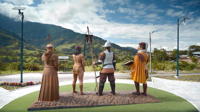 Vier Figuren stehen als Skulpturen in der Landschaft.