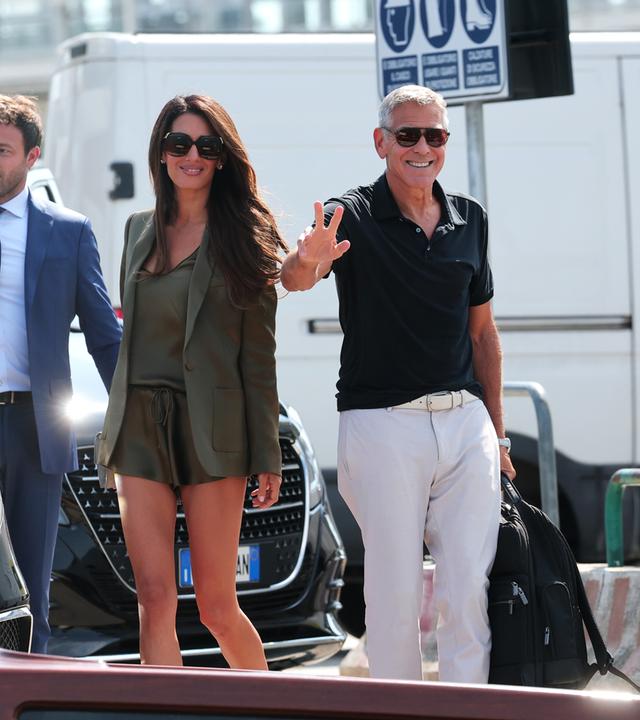 Das BIld zeigt Amal und George Clooney, die in eine Kamera lächeln und winken.