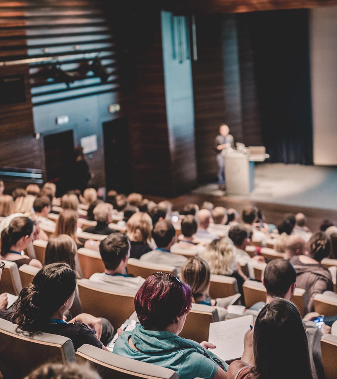 Blick über die Reihen eines voll besetzten Hörsaals während eines Vortrags mit Präsentation. Die Zuhörenden verfolgen die Inhalte konzentriert, was auf ein Seminar oder einen Workshop hindeutet. Solche Formate können auch von Finanzvertrieben genutzt werden, um Studierende anzusprechen.