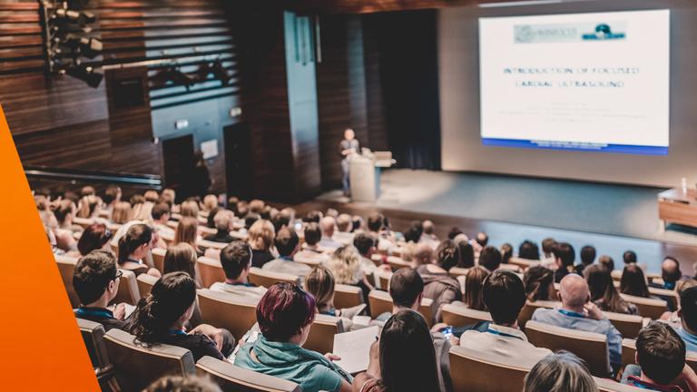 Blick über die Reihen eines voll besetzten Hörsaals während eines Vortrags mit Präsentation. Die Zuhörenden verfolgen die Inhalte konzentriert, was auf ein Seminar oder einen Workshop hindeutet. Solche Formate können auch von Finanzvertrieben genutzt werden, um Studierende anzusprechen.