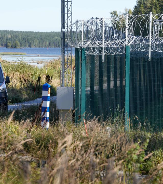 Ein Grenzzaun mit Stacheldraht. Links im Hintergund steht ein Polizeiauto