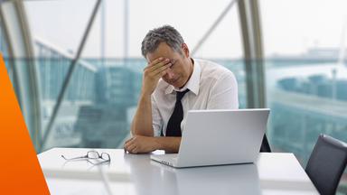 Ein Mann sitzt in einem modernen Büro an einem Tisch mit Laptop und wirkt nachdenklich und belastet. Er hat die Augen geschlossen oder gesenkt und stützt den Kopf mit der Hand. Auf dem Tisch liegt eine Brille, im Hintergrund sind große Fenster mit Blick nach draußen.