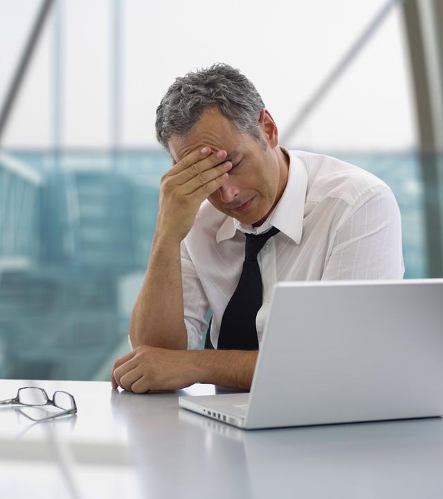 Ein Mann sitzt in einem modernen Büro an einem Tisch mit Laptop und wirkt nachdenklich und belastet. Er hat die Augen geschlossen oder gesenkt und stützt den Kopf mit der Hand. Auf dem Tisch liegt eine Brille, im Hintergrund sind große Fenster mit Blick nach draußen.
