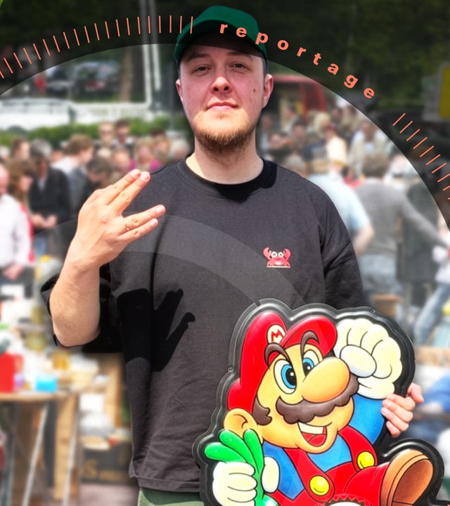 Jonas steht vor einem großen Auto und hält eine Wanddekoration von einer alten Videospielkonsole in der Hand.