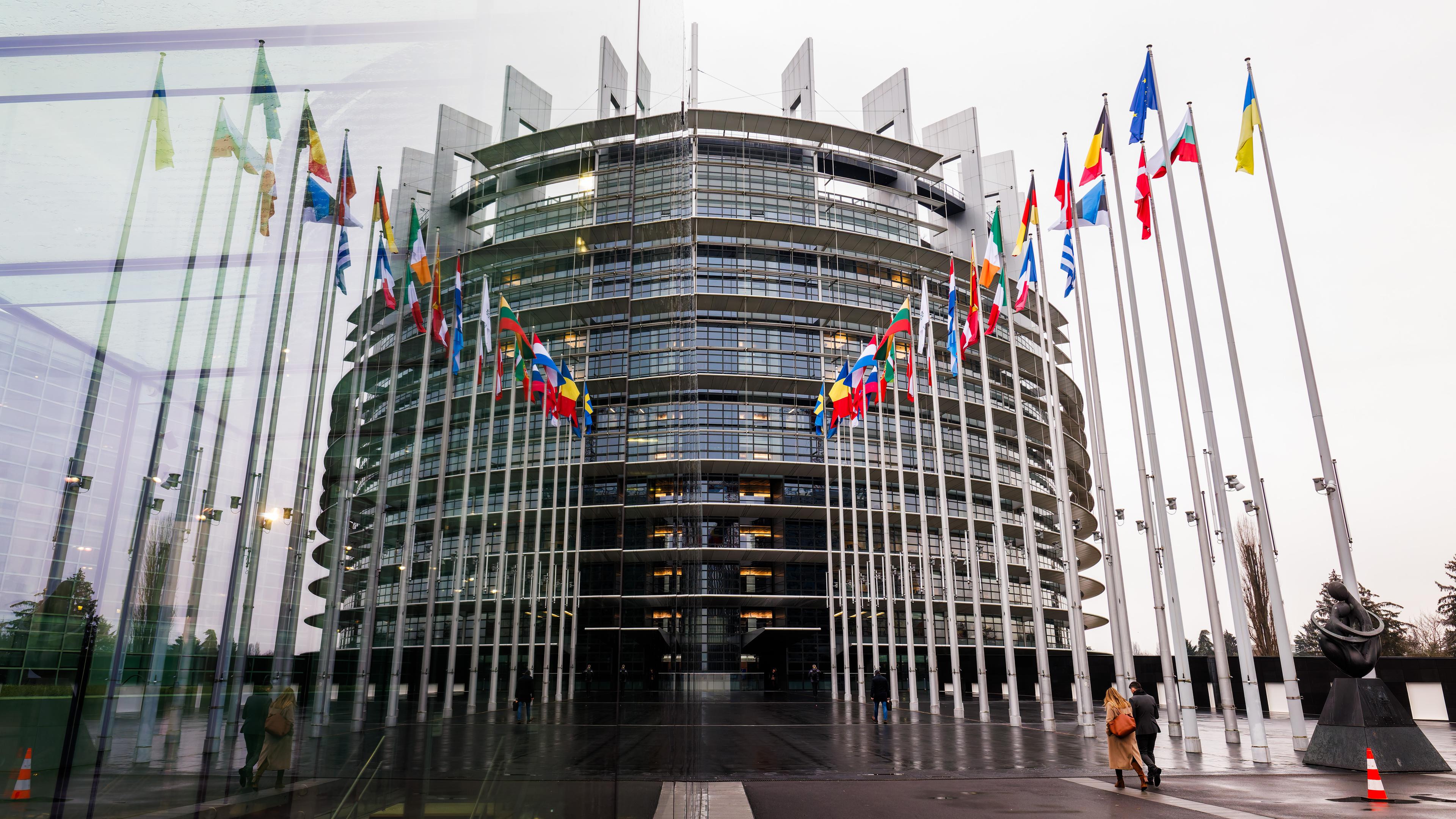 Die Fahnen der Mitgliedsstaaten der Europäischen Union und der Ukraine wehen vor dem Gebäude des Europäischen Parlaments und spiegeln sich dabei in einer gläsernen Wand.