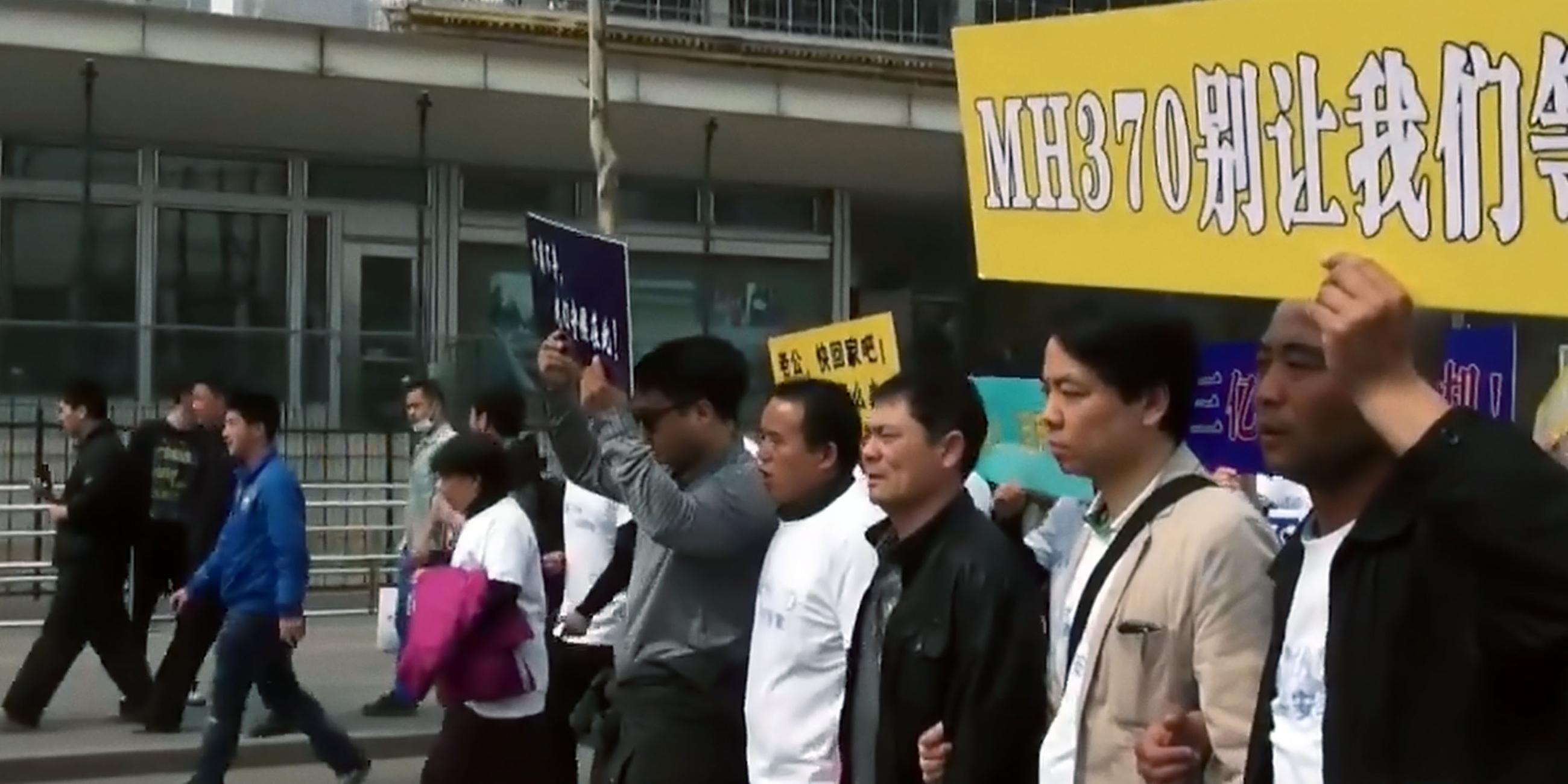 Eine Gruppe Menschen gehen in einer Reihe eine Straße entlang. Sie tragen ein Schild, auf dem in weißen Buchstaben auf gelbem Grund MH370 steht, daneben stehen chinesische Schriftzeichen.
