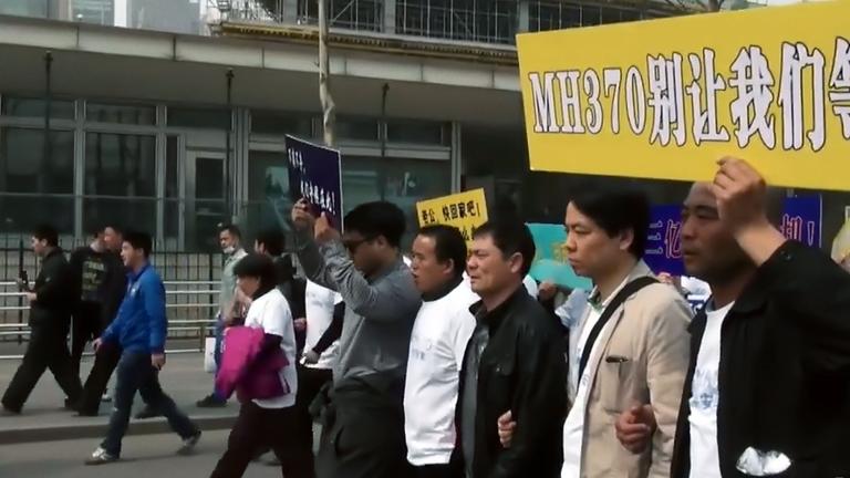 Eine Gruppe Menschen gehen in einer Reihe eine Straße entlang. Sie tragen ein Schild, auf dem in weißen Buchstaben auf gelbem Grund MH370 steht, daneben stehen chinesische Schriftzeichen.