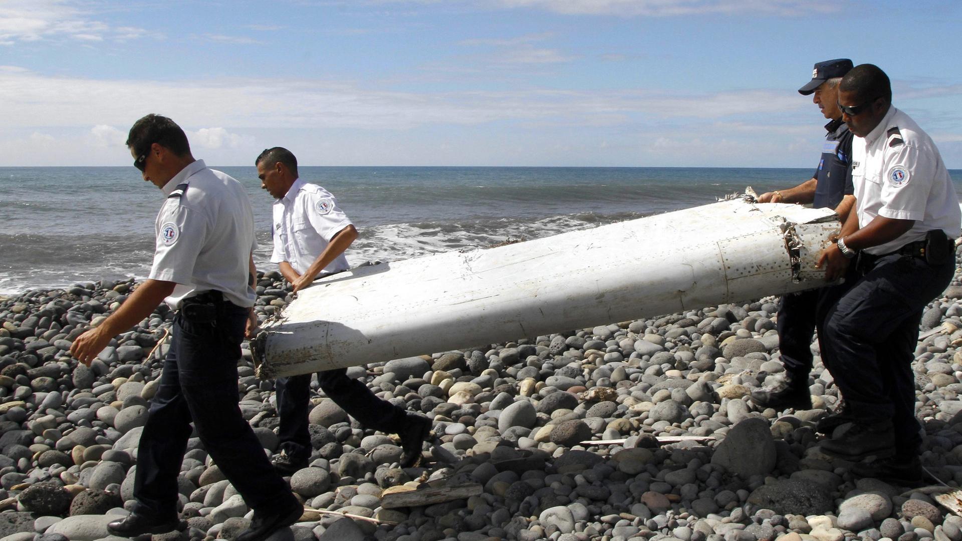 Vier Männer in weißen Hemden und blauen Hosen tragen ein etwa zwei Meter großes, weißes Trümmerteil über einen Steinstrand.
