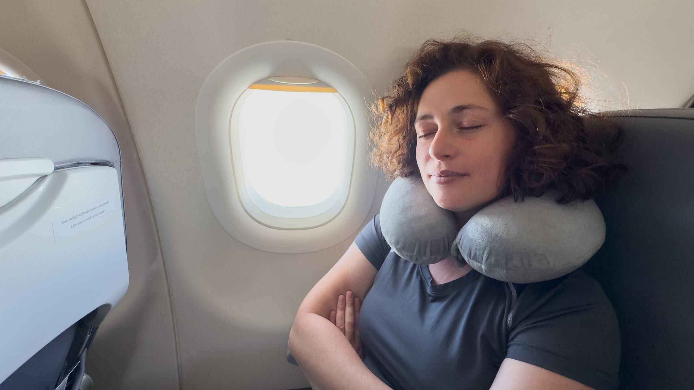 Eine Frau sitzt mit einem Nackenkissen im Flugzeug und hat die Augen geschlossen.