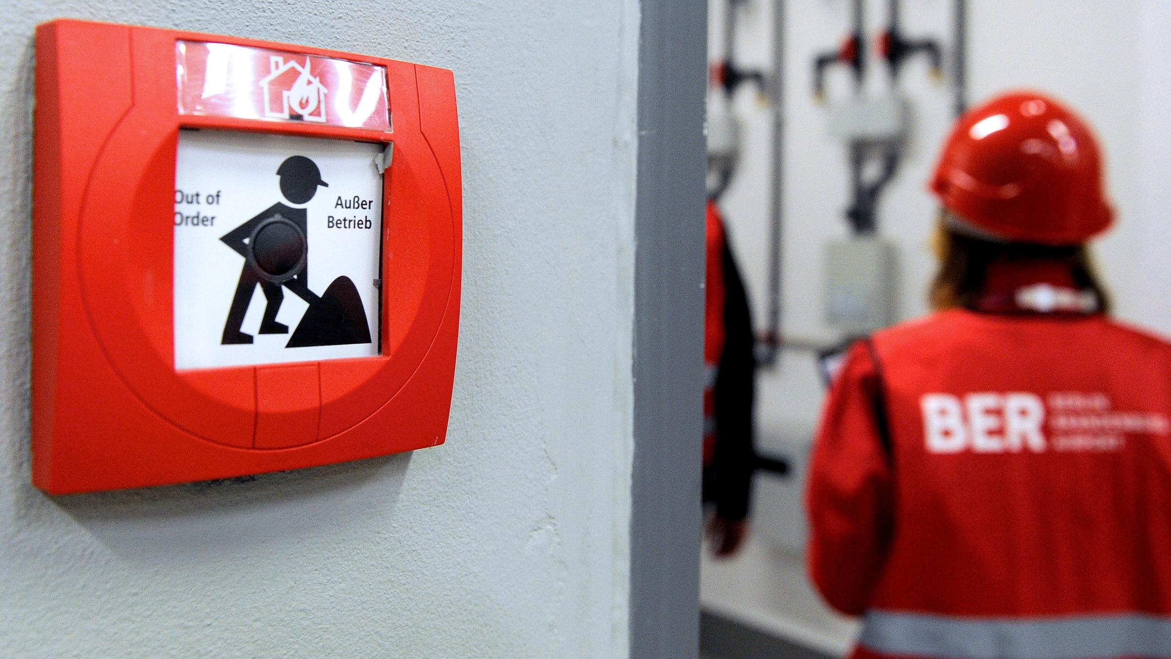 Archiv: Eine Frau mit Bauhelm und Warnweste läuft am 02.11.2012 auf dem Flughafen Berlin Brandenburg (Ber) in Schönefeld an einem Feuermelder vorbei