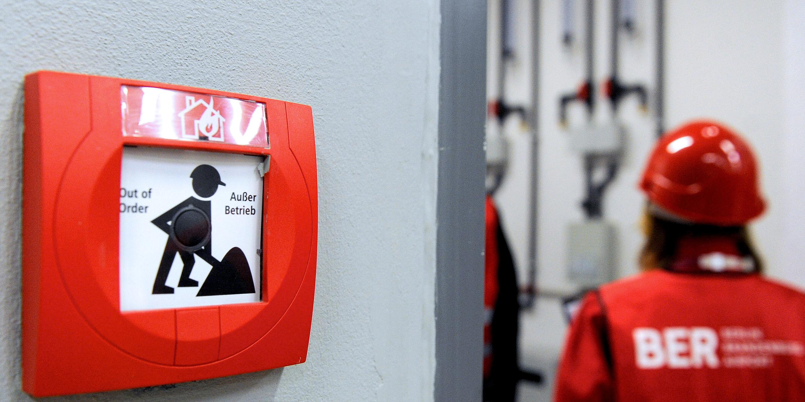 Archiv: Eine Frau mit Bauhelm und Warnweste läuft am 02.11.2012 auf dem Flughafen Berlin Brandenburg (Ber) in Schönefeld an einem Feuermelder vorbei