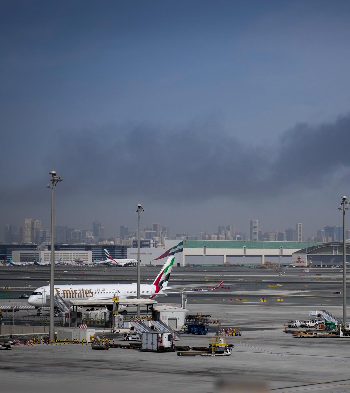 Eine Rauchwolke, verursacht durch einen iranischen Angriff, ist im Hintergrund zu sehen, während Flugzeuge der Emirates nach der Schließung des Flughafens auf dem Dubai International Airport geparkt sind.