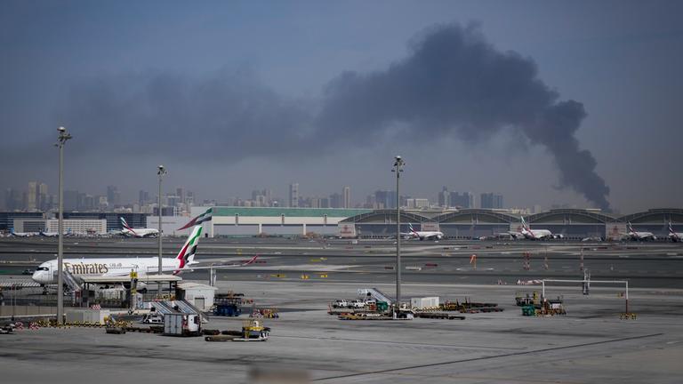 Eine Rauchwolke, verursacht durch einen iranischen Angriff, ist im Hintergrund zu sehen, während Flugzeuge der Emirates nach der Schließung des Flughafens auf dem Dubai International Airport geparkt sind.