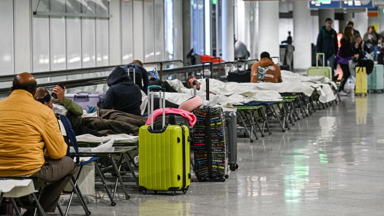 Menschen warten am Donnerstag Abend auf Feldbetten am Flughafen in München. Wegen Drohnensichtungen war der Flugbetrieb eingestellt worden. Zahlreiche Flüge fiehlen aus. 