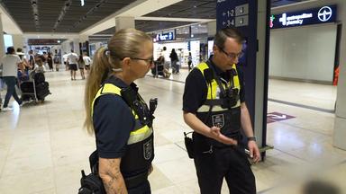Sicherheitskräfte in Dienstkleidung stehen in einem Terminal des Flughafens Berlin Brandenburg. Im Hintergrund bewegen sich Reisende durch die Halle.