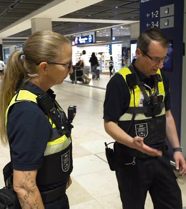 Sicherheitskräfte in Dienstkleidung stehen in einem Terminal des Flughafens Berlin Brandenburg. Im Hintergrund bewegen sich Reisende durch die Halle.