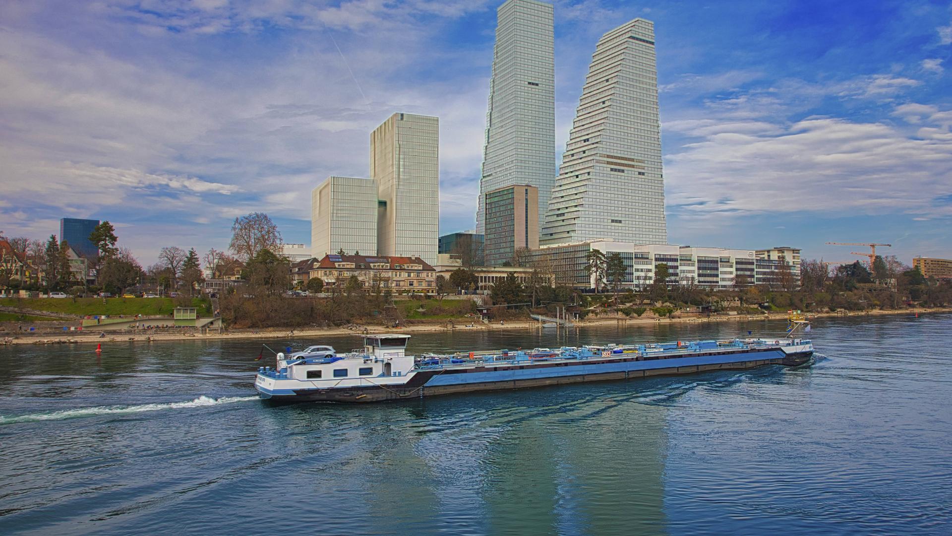 Ein Frachtschiff fährt auf dem Fluss in Basel, flankiert von modernen Hochhausbauten des Pharmakonzerns Roche