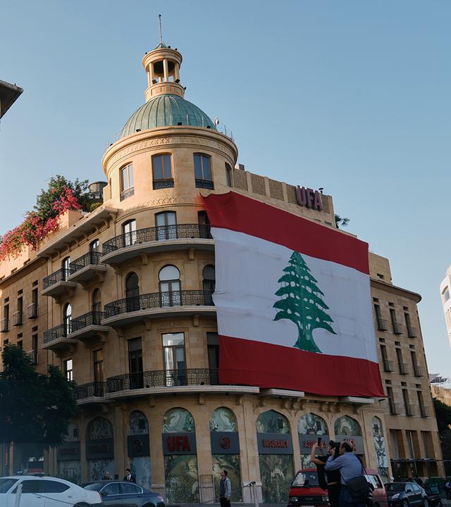 Zu sehen ist eine Straße und besonders zwei Gebäude mit großen libanesischen Flaggen, die einen Großteil der Gebäude-Fronten verdecken.