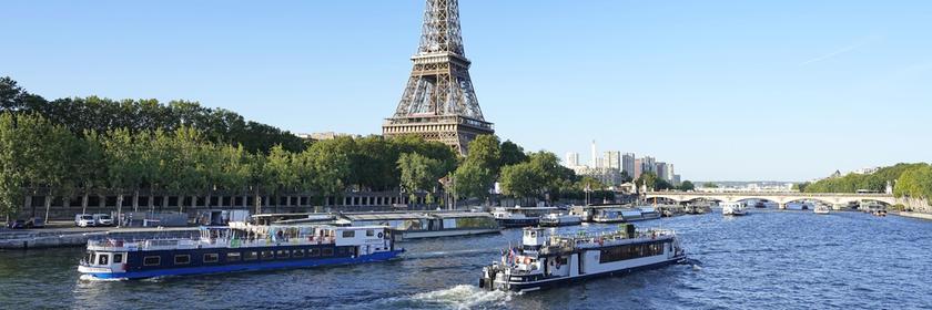Der Eiffelturm ist in der Mitte des Bildes. Im Vordergrund sieht man die Seine auf der zwei Schiffe fahren. Der Himmel ist blau.