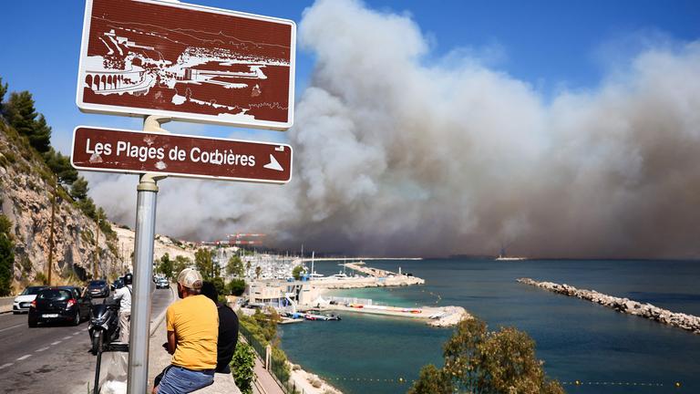 Menschen schauen zu in der Nähe von Marseille zu, während im Hintergrund der Rauch eines Waldbrandes wütet.