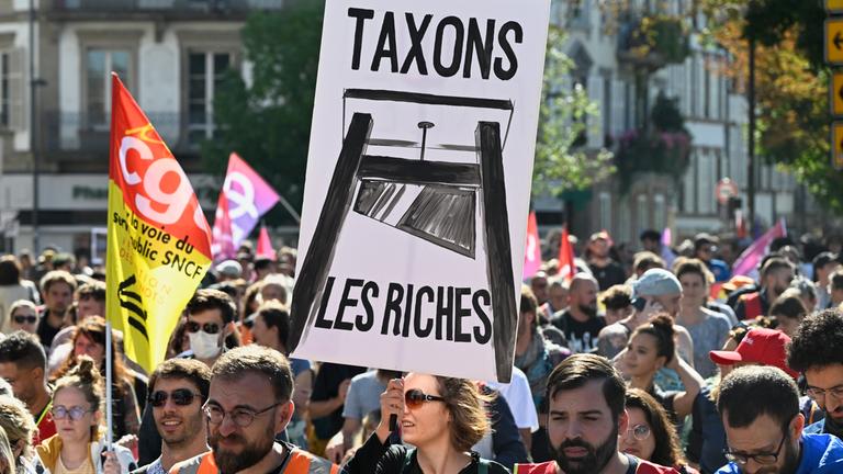 Frankreich, Straßburg: Demonstranten ziehen durch eine Straße. Eine Frau hält ein Plakat mit der Aufschrift "Taxons les Riches" in den Händen.