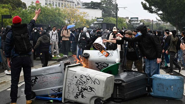In Nantes protestieren Menschen. Im Vordergrund sind mehrere mit Graffitis versehene Mülleimer.