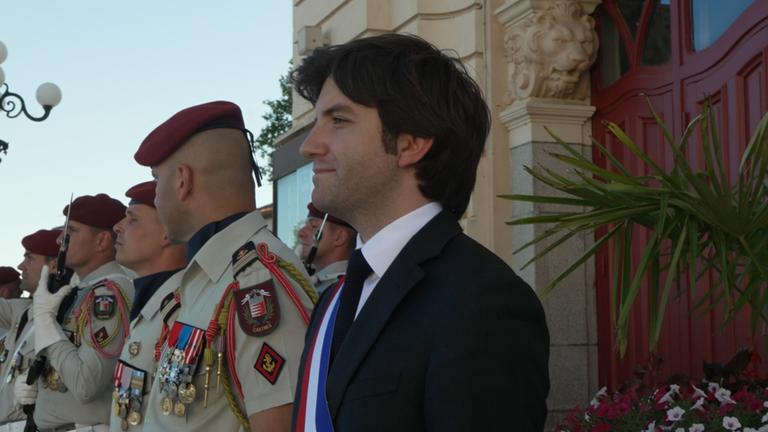 Florian Azéma, Bürgermeister der französischen Stadt Castres, steht vor einem Haus mit ein paar Männern in Militäruniform.
