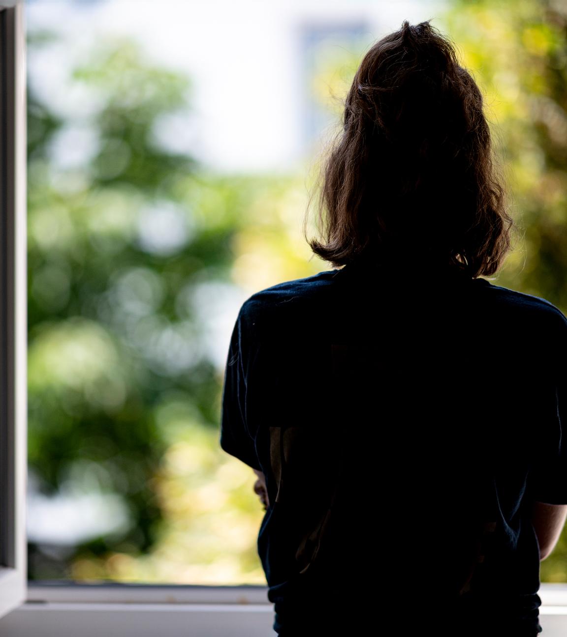 Rückansicht einer jungen Frau mit zusammengebundenen Haaren, die aus einem geöffneten Fenster schaut.