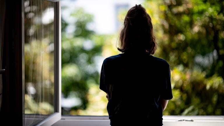 Rückansicht einer jungen Frau mit zusammengebundenen Haaren, die aus einem geöffneten Fenster schaut.