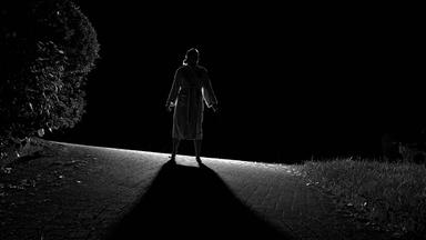 Eine Frau steht bei Nacht auf einer Anhöhe mit dem Rücken zum Betrachter und wird von vorne angeleuchtet, so dass sie als Schattenriss zu sehen ist.
