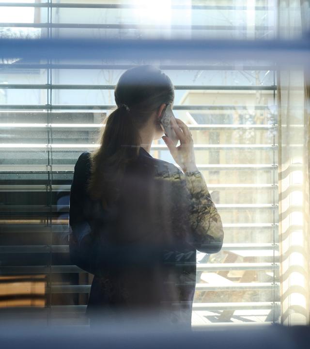 Symbolbild:Eine Frau telefoniert während der Arbeit mit einem Smartphone und blickt aus einem Fenster mit Jalousien. 