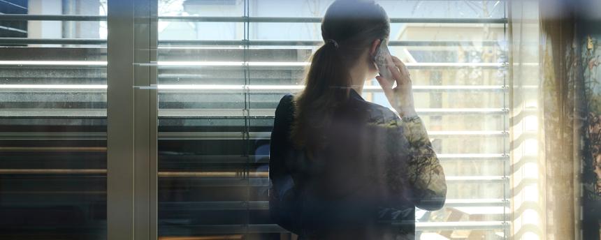 Symbolbild:Eine Frau telefoniert während der Arbeit mit einem Smartphone und blickt aus einem Fenster mit Jalousien. 
