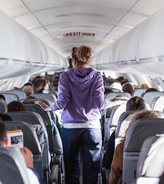 Der Innenraum eines Flugzeugs mit Passagieren auf den Sitzen und einer Reisenden, die durch den Gang geht.