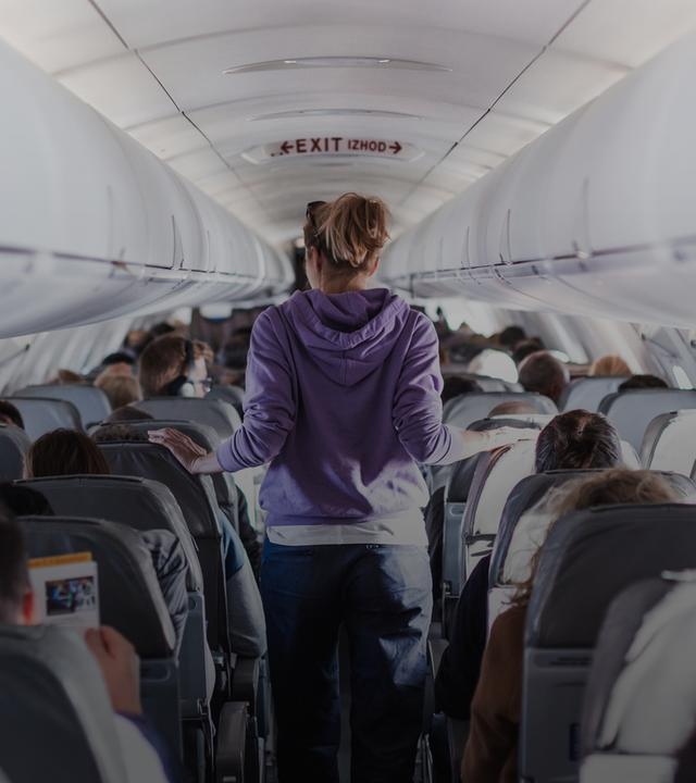 Der Innenraum eines Flugzeugs mit Passagieren auf den Sitzen und einer Reisenden, die durch den Gang geht.