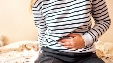Eine Frau in einem gestreiften Langarmshirt sitzt auf dem Bett und hält sich vor Bauchschmerzen den Bauch.
