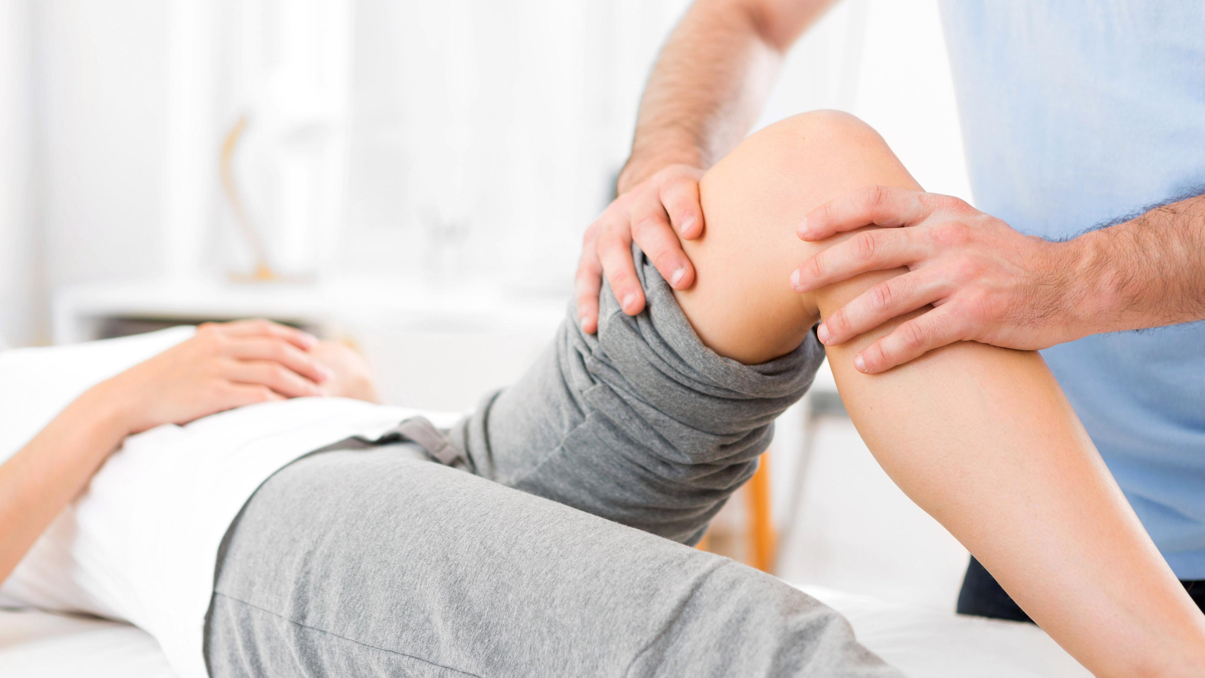 Eine Frau liegt auf dem Rücken. Sie trägt Sportkleidung und ihr linkes Bein ist angewinkelt. Ein Mann untersucht ihr Knie auf Verletzungen.