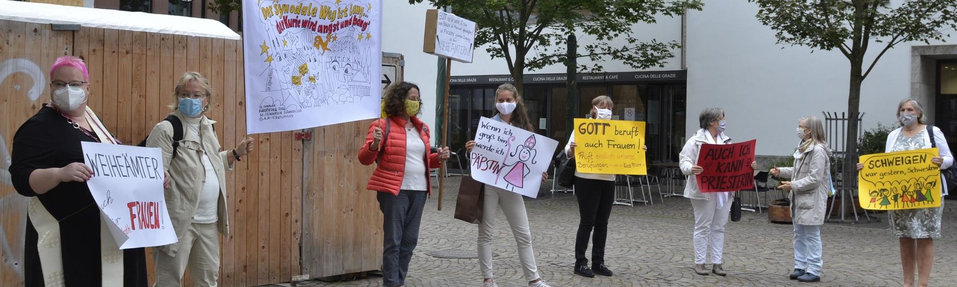 Eine Gruppe von Frauen steht auf einem Platz und hält ihre bunten Protestschilder in den Händen.
