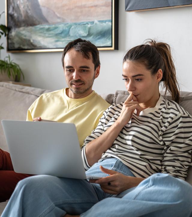 Eine Frau und ein Mann sitzen auf einer Couch, beiden blicken auf einen auf einen aufgeklappten Laptop.