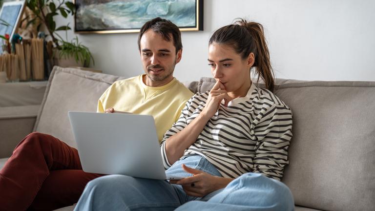 Eine Frau und ein Mann sitzen auf einer Couch, beiden blicken auf einen auf einen aufgeklappten Laptop.