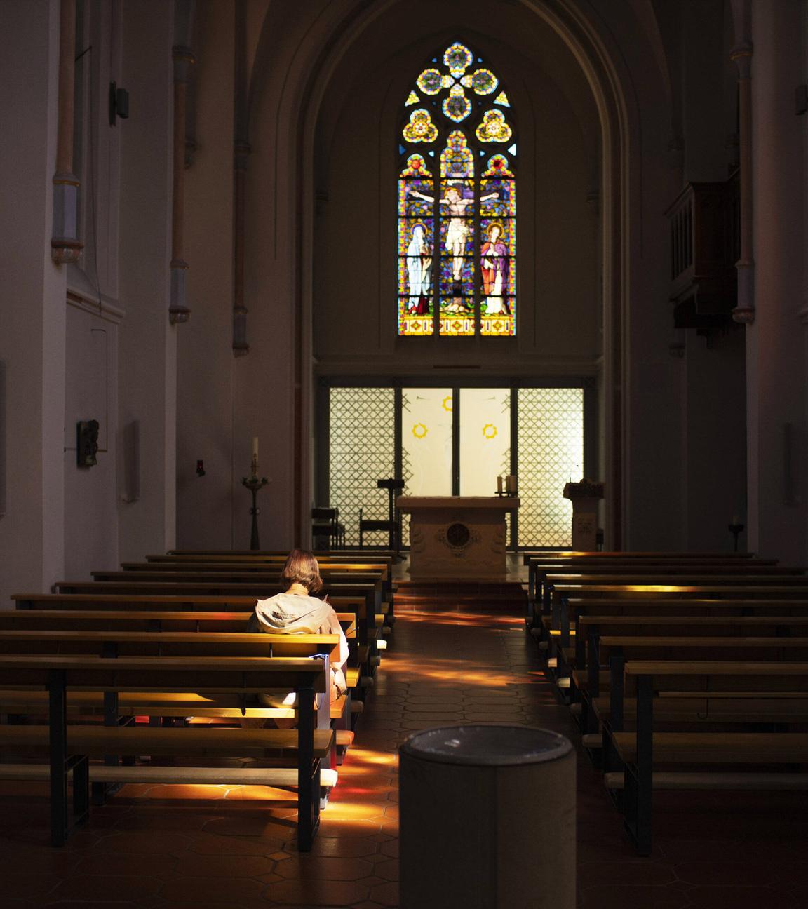 Rückansicht einer Frau, die alleine in einer Kirchenbank in einer sonst leeren alten Kirche mit einem großen Buntglasfenster über dem Altar sitzt.