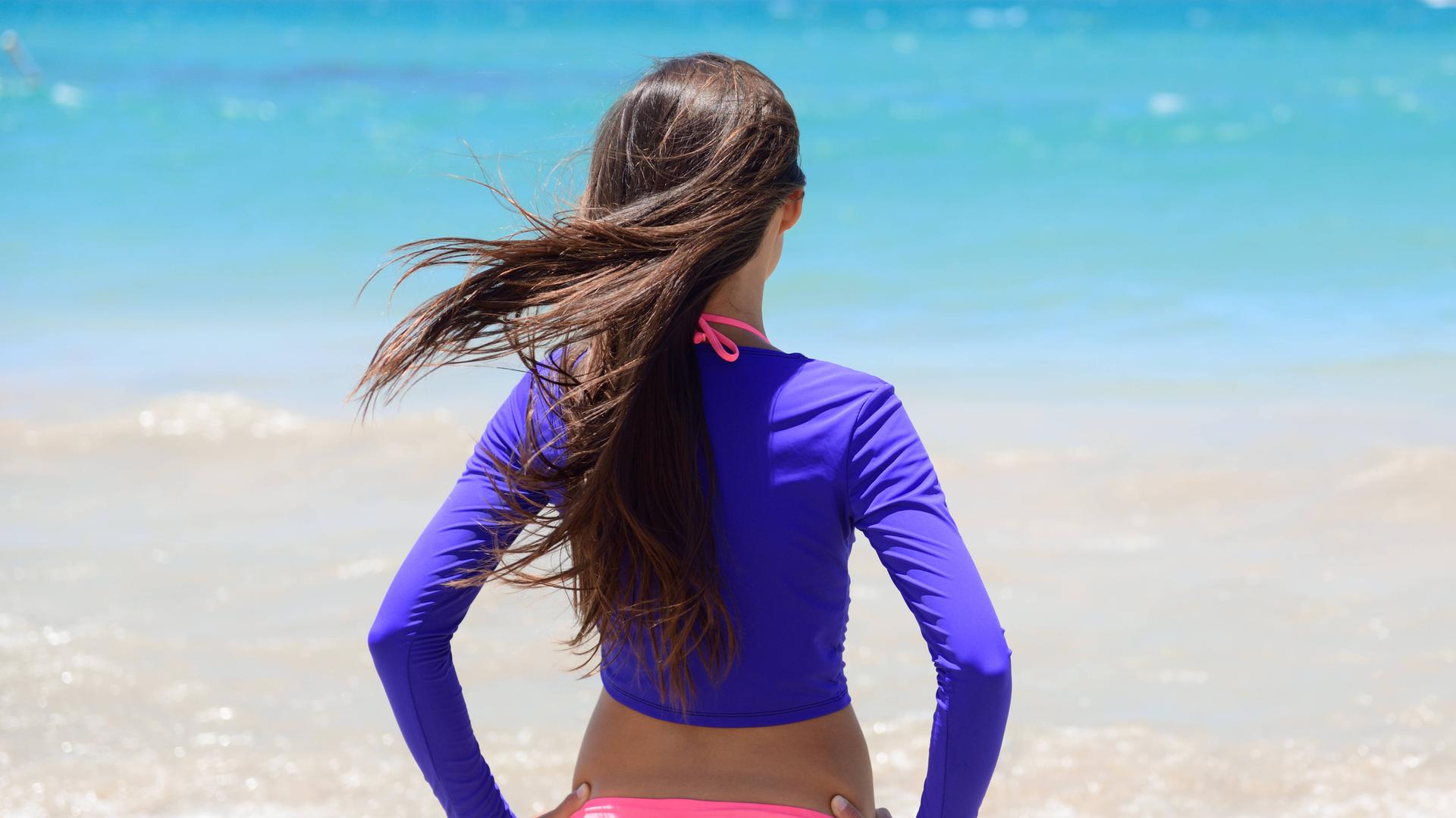 Ein Frau im Bikini mit einem langärmeligen T-Shirt als UV-Schutz steht am Strand und blickt auf das Meer.