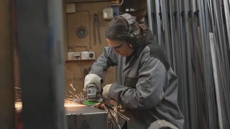 Die Ofenbauerin Laura Hack geht ihrer handwerklichen Tätigkeit nach. Sie trägt Schutzklamotten und eine Schutzbrille.