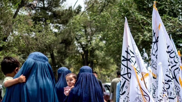 Afghanische Frauen in Burkas gehen am Vorabend des vierten Jahrestages der Machtübernahme der Taliban im Jahr 2021 am 14. 8. 2025 in Kabul an Taliban-Flaggen vorbei. 