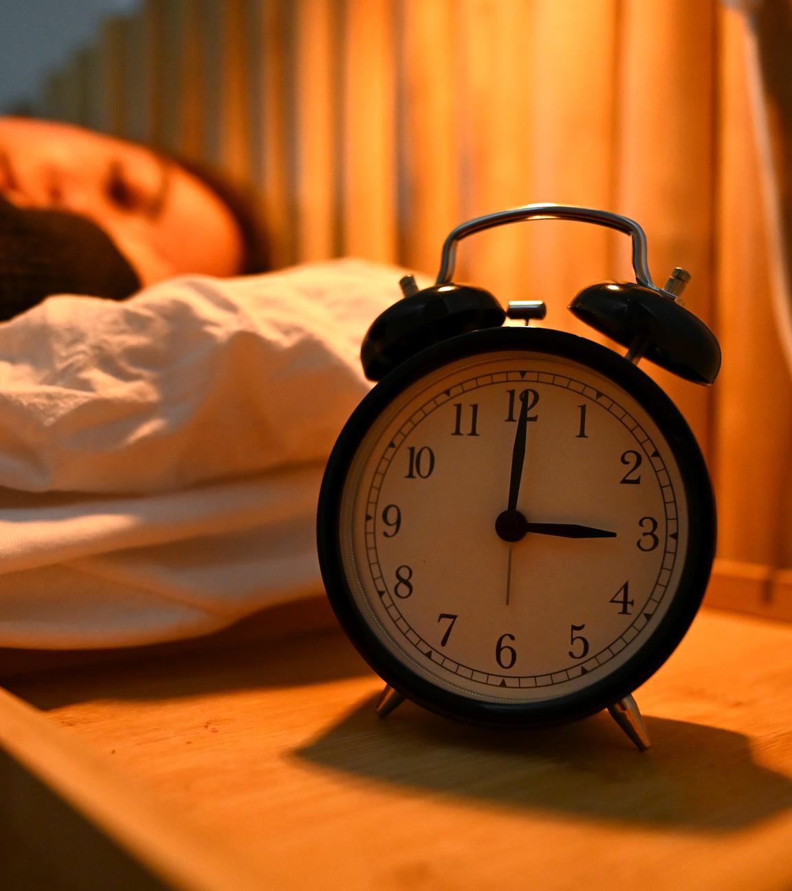 Ein Wecker steht neben einem Bett, in dem eine Frau schläft.